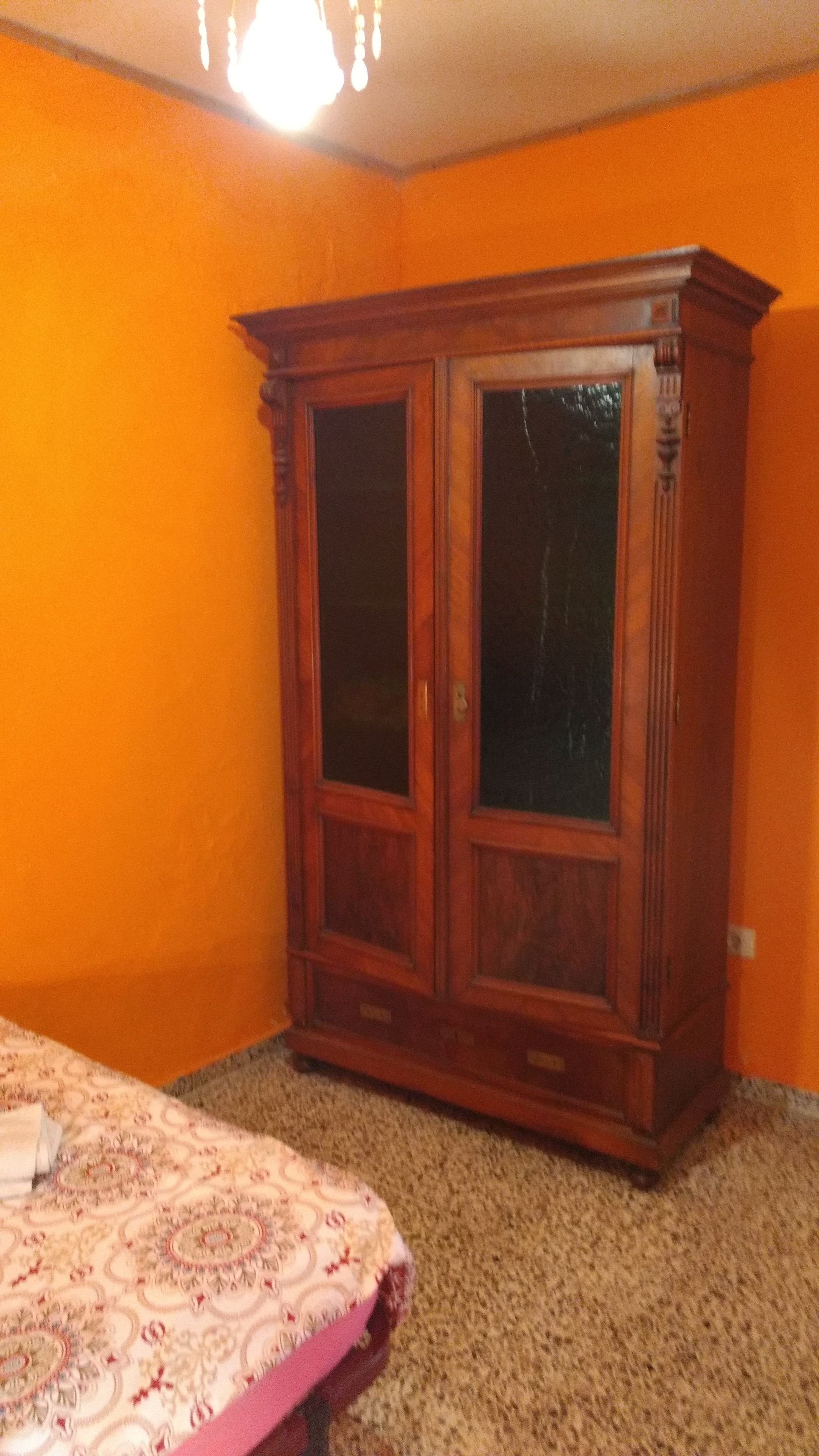 Una habitación con un armario grande de madera frente a una pared naranja, suelo alfombrado y una colcha estampada.