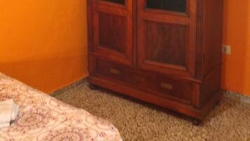 Una habitación con un armario grande de madera frente a una pared naranja, suelo alfombrado y una colcha estampada.