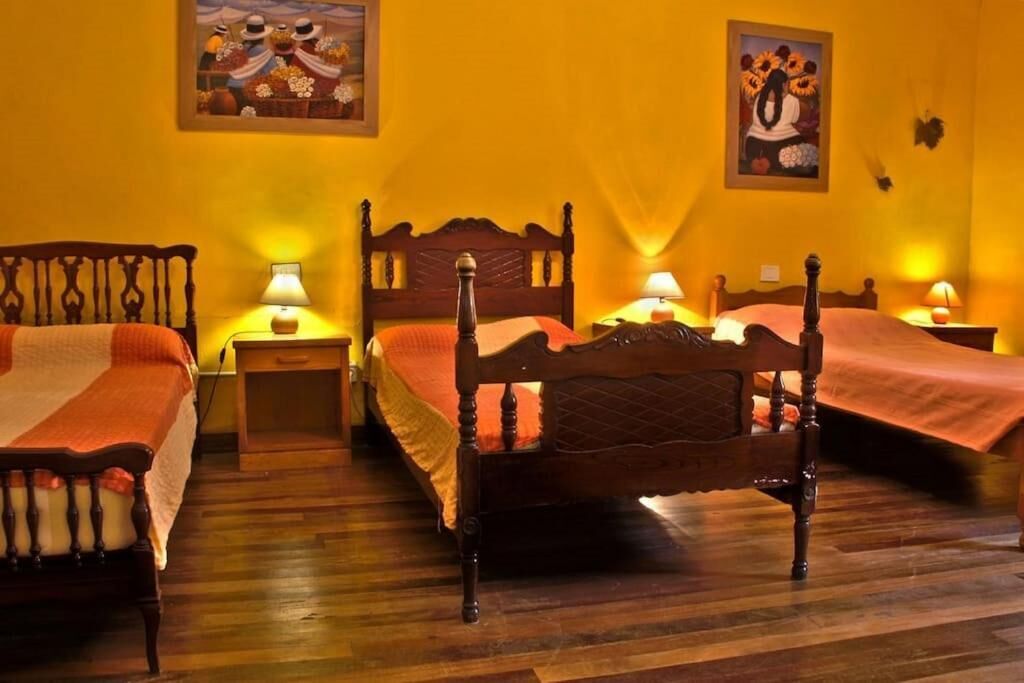 Een kamer met drie houten bedden, elk voorzien van een nachtkastje en een lamp. De muren zijn geel geschilderd, en er hangen twee schilderijen aan de muur.