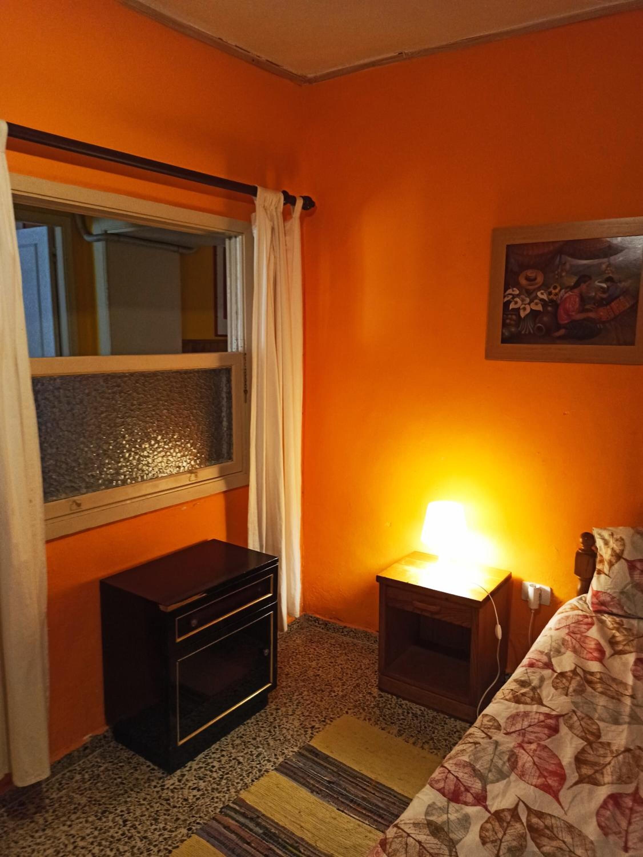 Quarto com parede laranja, janela com cortinas, pequena mesa de cabeceira de madeira com abajur, cama com colcha estampada e quadro na parede.