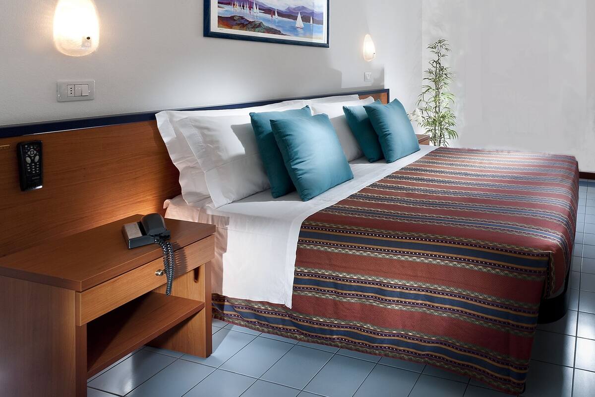 A hotel room with a wooden headboard, a bed with a striped bedspread, a bedside table with a phone, and a painting on the wall.