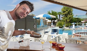 Ein Mann gießt ein Getränk in ein Glas an einer Poolbar mit Blick auf Pool und Liegestühle.