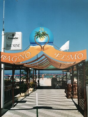 Une promenade en bord de mer mène à un bar de plage appelé « BAGNO ELMO », dont l’enseigne, sur le thème tropical, représente des palmiers et un soleil.
