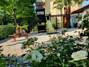 Ein Spielplatz mit Rutsche und Schaukeln, umgeben von Grünflächen und einem Gebäude im Hintergrund.