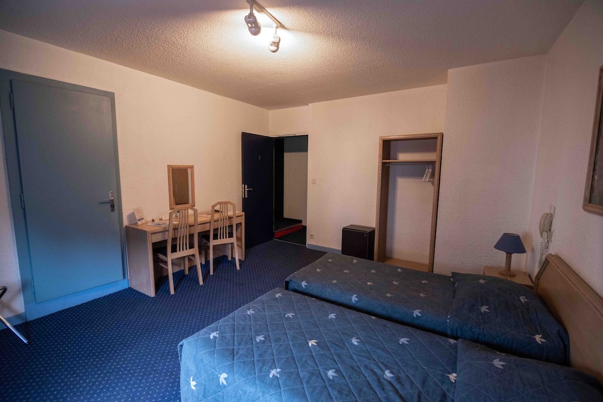 A room with two beds, a desk, a chair, a wardrobe, and a small table with a lamp.