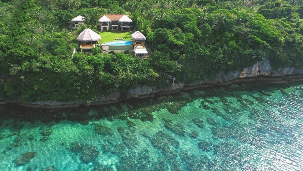 An aerial view of a resort with multiple thatched-roof buildings, a swimming pool, and lush greenery surrounding a coastal area.