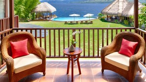 A balcony with wicker chairs, a small table, and a view of a pool and mountains.