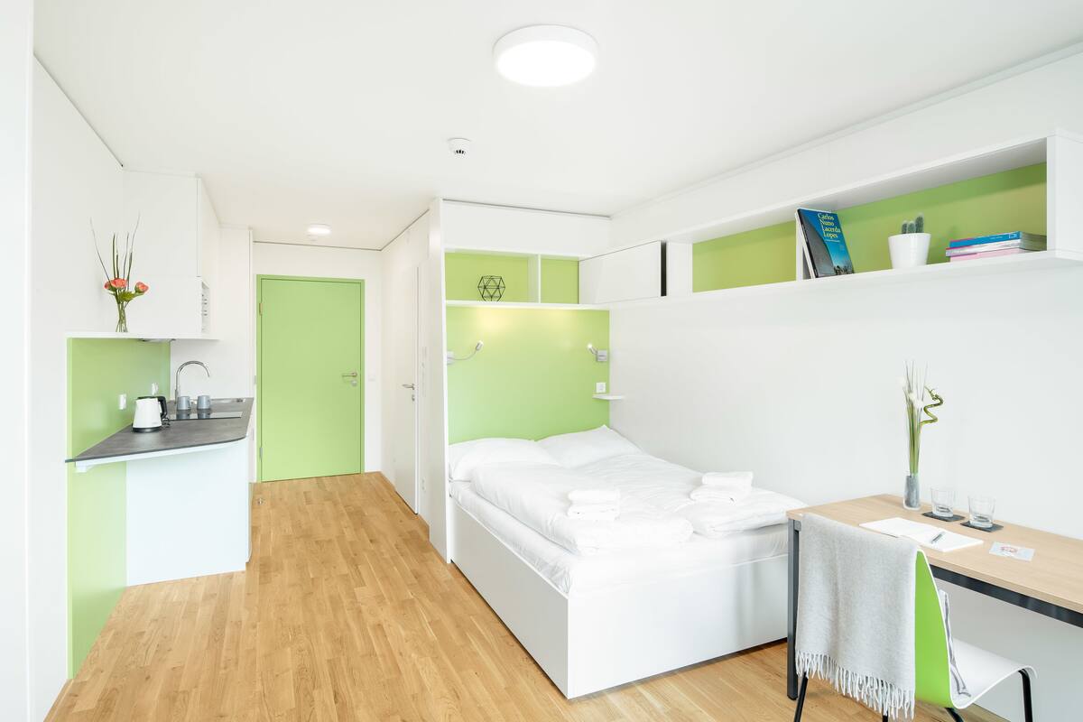 A modern, compact room with a bed, a desk, and a kitchenette area.