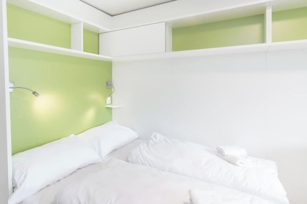 A modern hotel room with a bed, white bedding, a green accent wall, and a nightstand with a lamp.