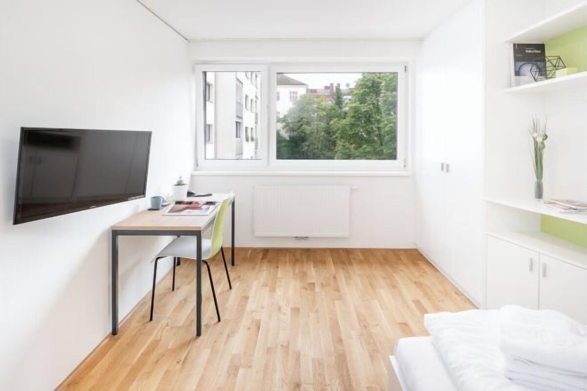 A modern, compact room with a wooden floor, a bed, a desk with a chair, a mounted TV, and a window with a view of buildings and trees.