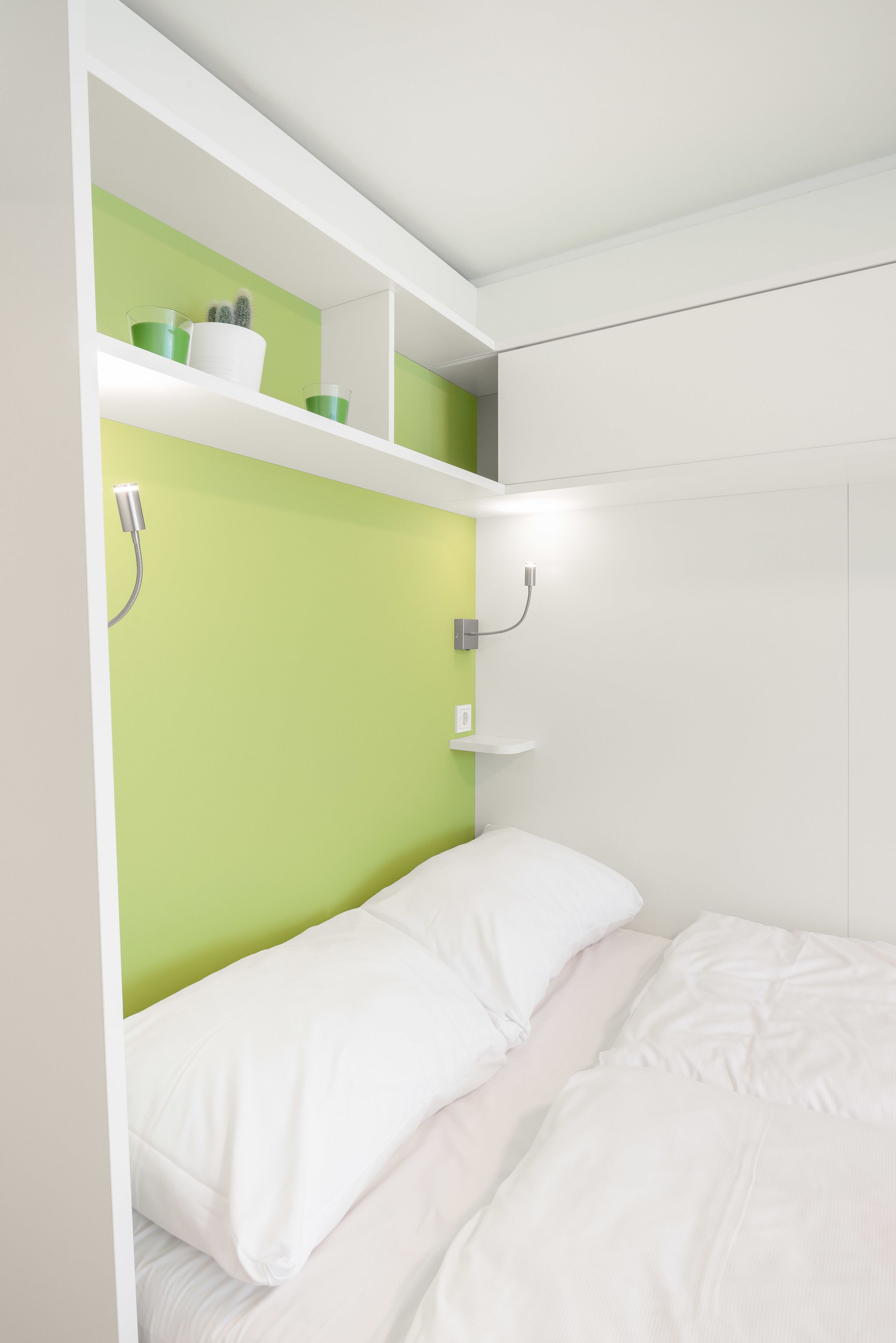 Un dormitorio con una pared verde decorativa, ropa de cama blanca y una estantería con objetos ornamentales.