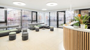 A modern reception area with a wooden reception desk, seating arrangements, and large windows offering views of the street. - myNext - Hotel Leo (Vienna)