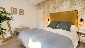 Ein Schlafzimmer mit einem Kopfende aus Steinoptik, einem großen Bett mit grauer Bettwäsche, einem Nachttisch mit Lampe und einem Spiegel mit goldener Rahmung.