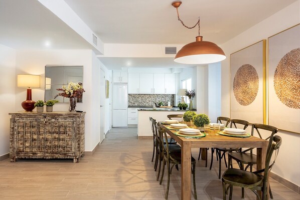 Une cuisine moderne avec des meubles blancs, un coin repas doté d’une table et de chaises en bois, et une lampe décorative.