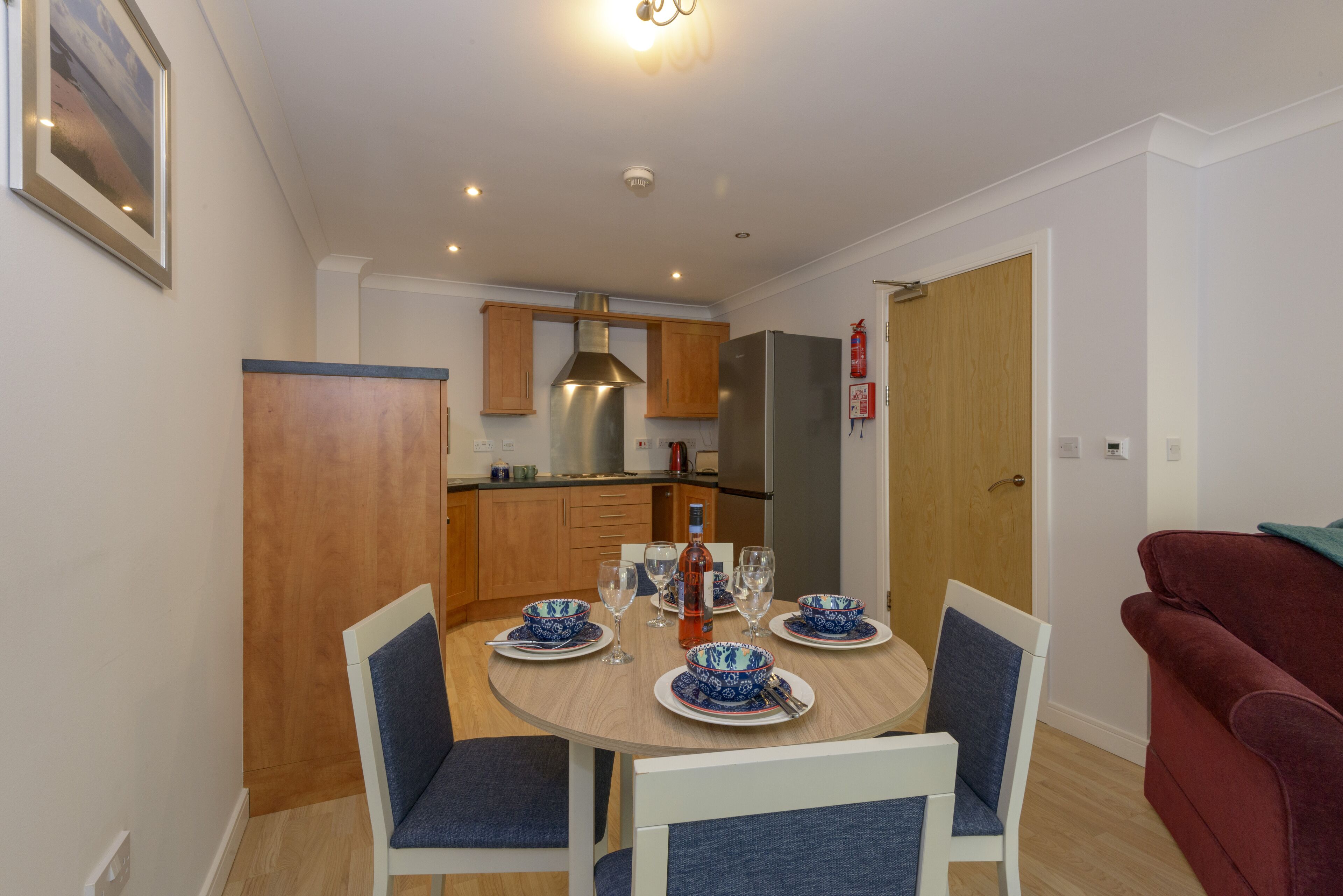A dining area with a round table set for four, surrounded by chairs. A kitchen with wooden cabinets and a stainless steel range hood is visible in the background.