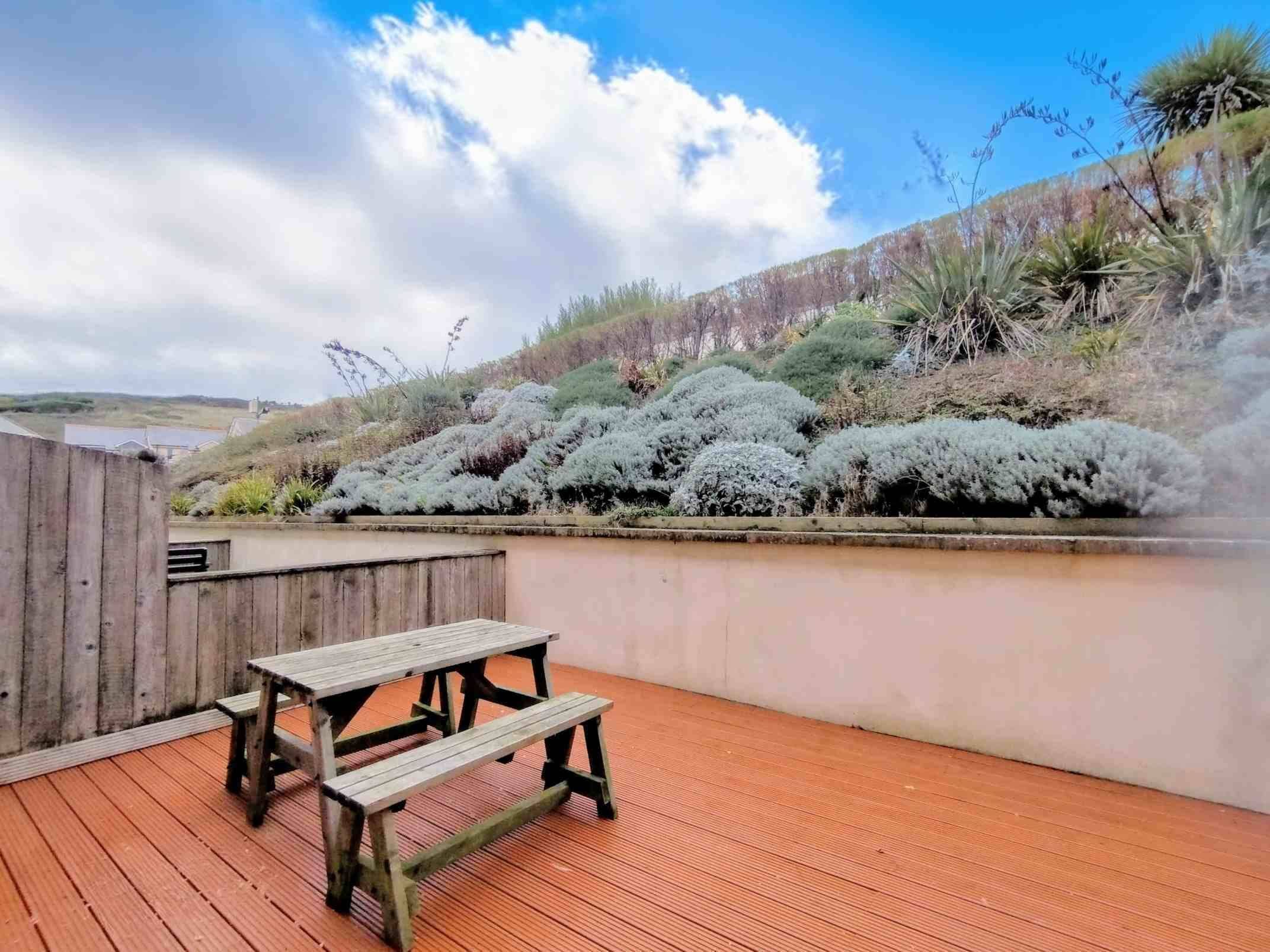En träaltan med ett picknickbord, omgiven av en låg mur och tät buskvegetation.