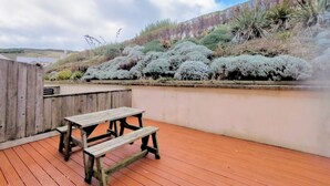 A wooden deck with a picnic table and benches, surrounded by a low wall and dense shrubbery. - Waves Apartments (Newquay)