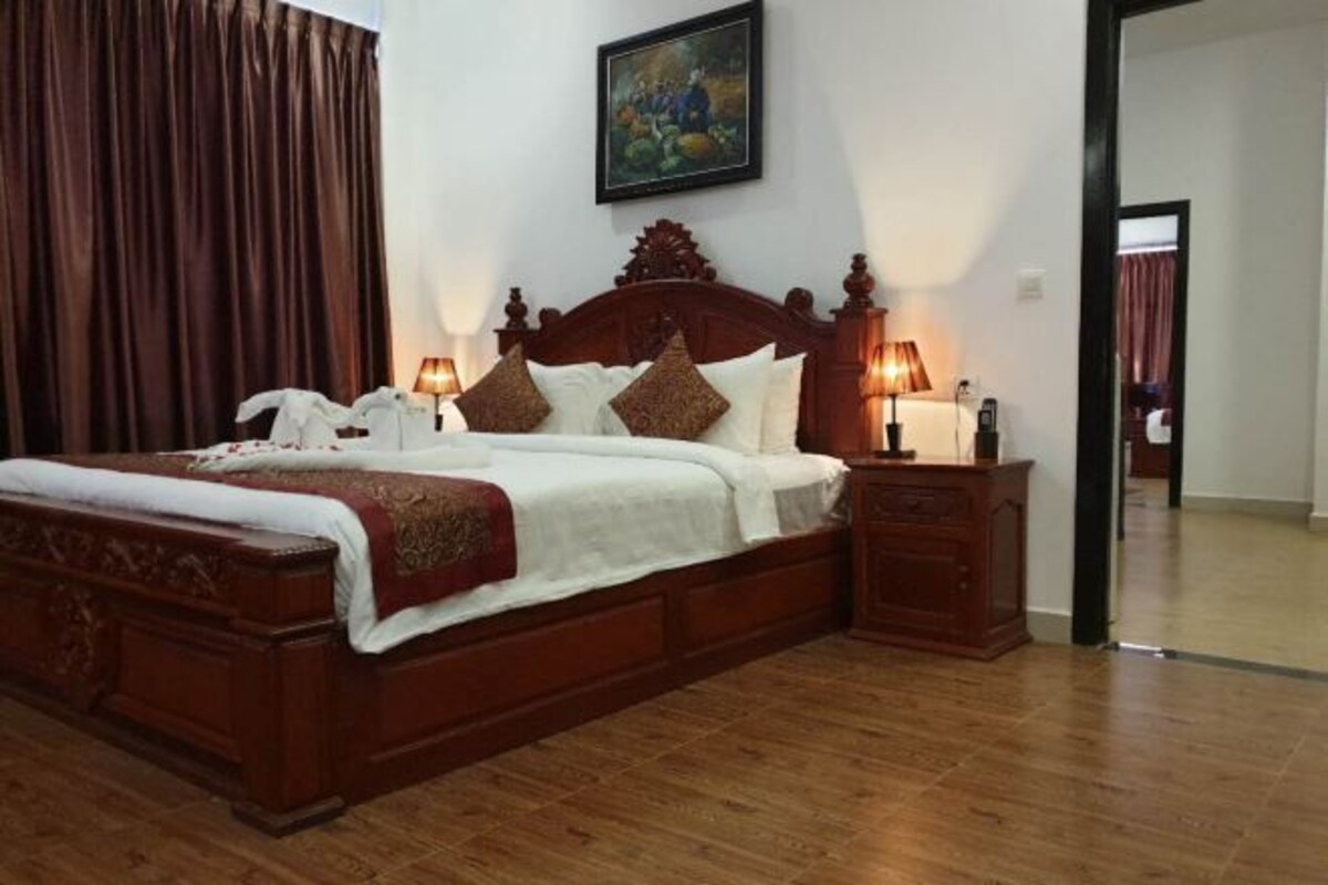 Una cama con dosel de madera, ropa de cama blanca y almohadas decorativas, una mesita de noche con lámpara y un cuadro en la pared.