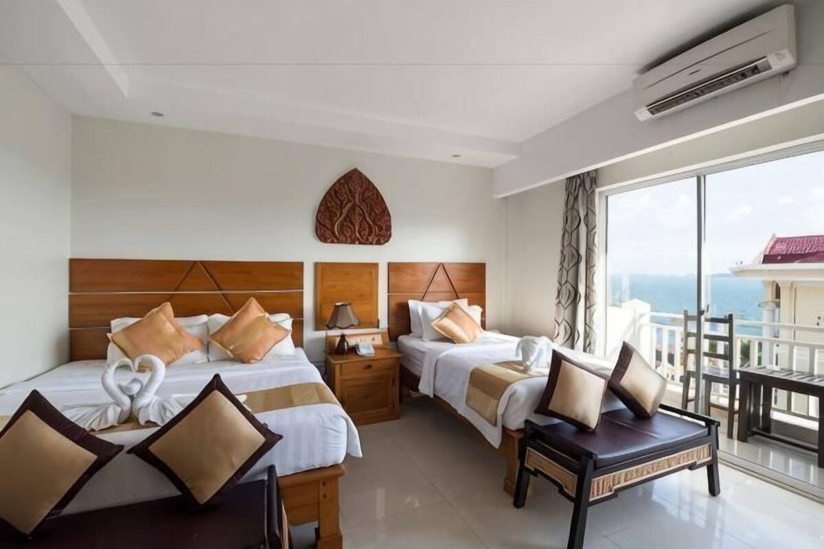 Une chambre d’hôtel avec deux lits, une tête de lit en bois, une table de chevet, un bureau, une chaise et un balcon avec vue sur la mer.