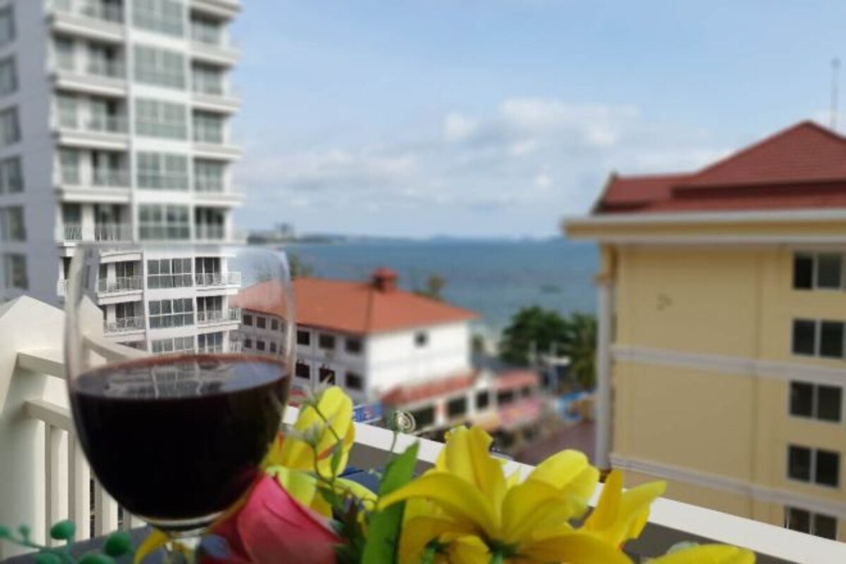 Un balcón con una copa de vino tinto y flores amarillas, con vistas a una ciudad costera con edificios y un cielo despejado.