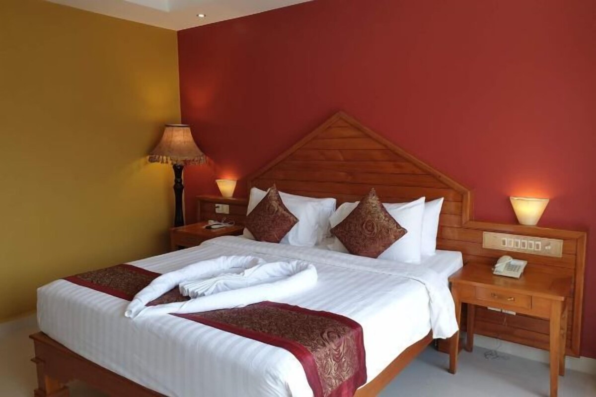 Una cama bien tendida con sábanas blancas y un plaid rojo, dos lámparas de noche y una mesita de noche de madera con un teléfono.