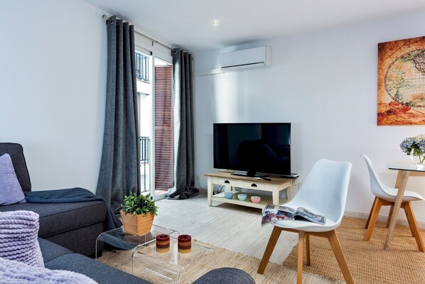 A modern living room with a sofa, a coffee table with a plant, a TV, and a dining area with chairs. - Genteel Home Albareda II (Sevilla)