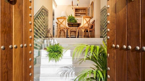 Una escalera conduce a un comedor con una araña, puertas de madera y plantas en macetas.
