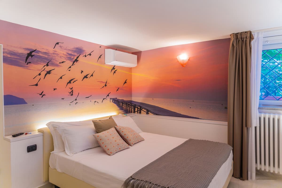 A hotel room with a bed, a nightstand, a wall-mounted air conditioner, a window with curtains, and a sunset mural on the wall.