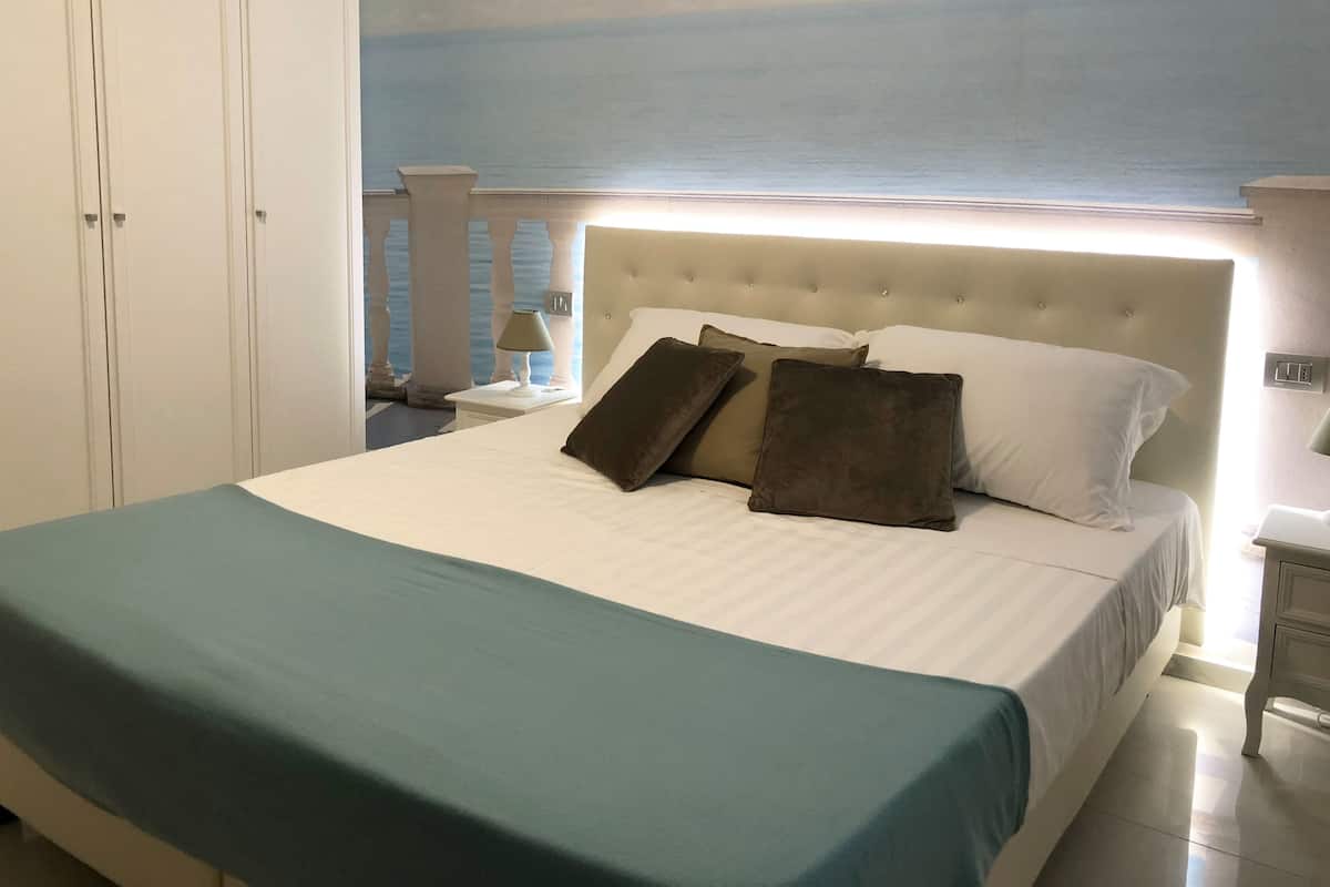A bedroom with a bed, a nightstand, a lamp, and a view of the sea through a window.