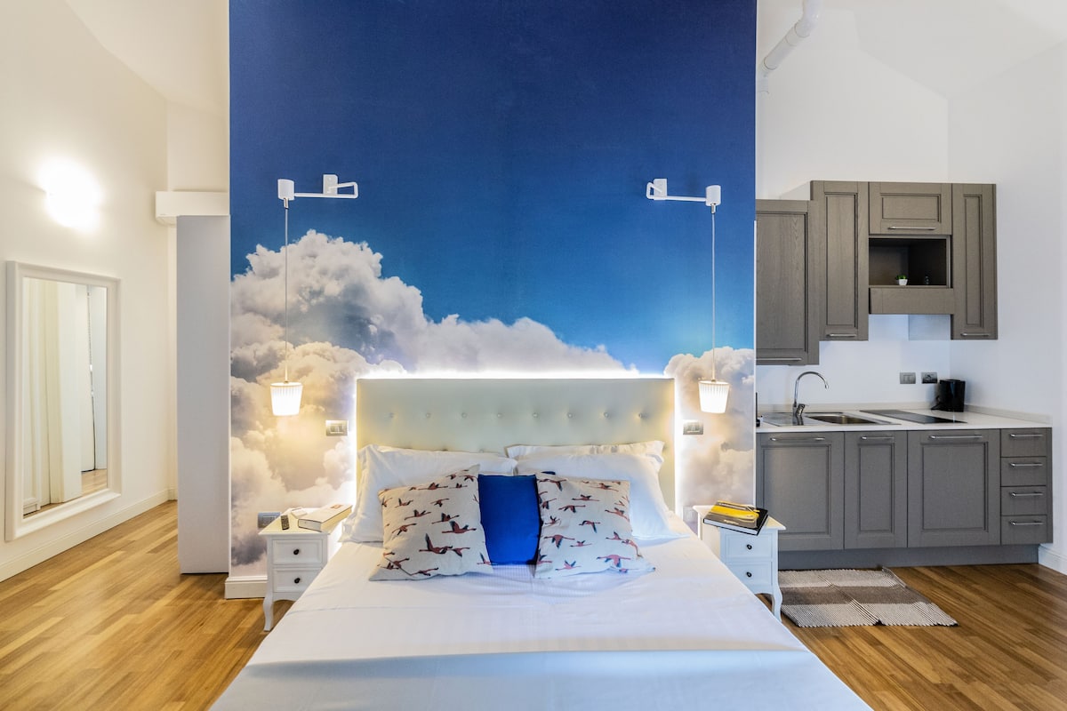 A bedroom with a bed, bedside tables, a nightstand, a wall mural, a window, and a kitchenette.