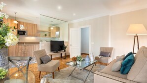 A modern living room with a sofa, chairs, a glass coffee table, and a TV. There is a kitchen area with wooden cabinets and a microwave. - Genteel Home Tomas Heredia (Malaga)