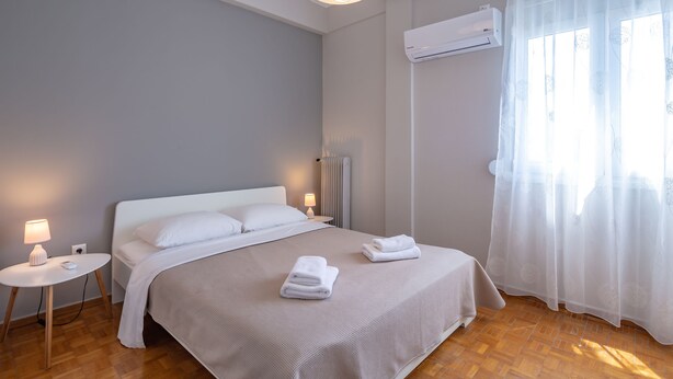 A modern bedroom with a bed, bedside lamps, an air conditioning unit, and a window with sheer curtains. - Artemis Deluxe Athens City Center Apartment (Athens)