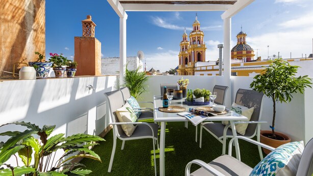 Une terrasse extérieure avec une table et des chaises blanches, entourée de plantes en pot et offrant une vue sur une église dotée de deux clochers.