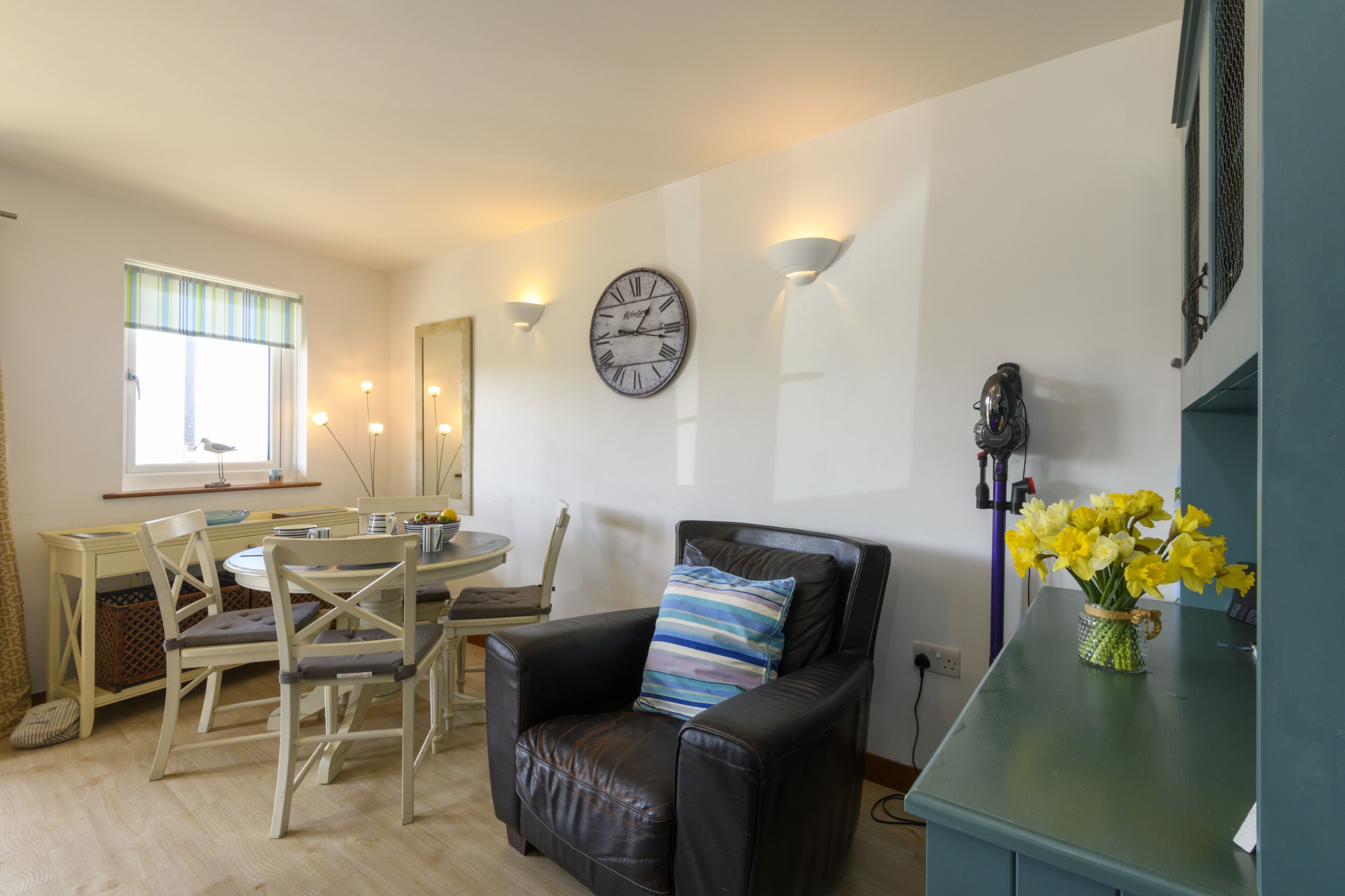 A living room with a dining area, a black leather sofa, a round table, and a wall clock.