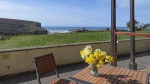 Eine Terrasse mit Tisch und Stühlen, eine Vase mit gelben Blumen und Blick auf das Meer und die Klippen.