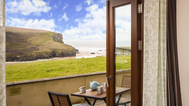 Ein Balkon mit Tisch und Stühlen, der auf eine Wiese und das Meer mit einer Klippe im Hintergrund blickt.