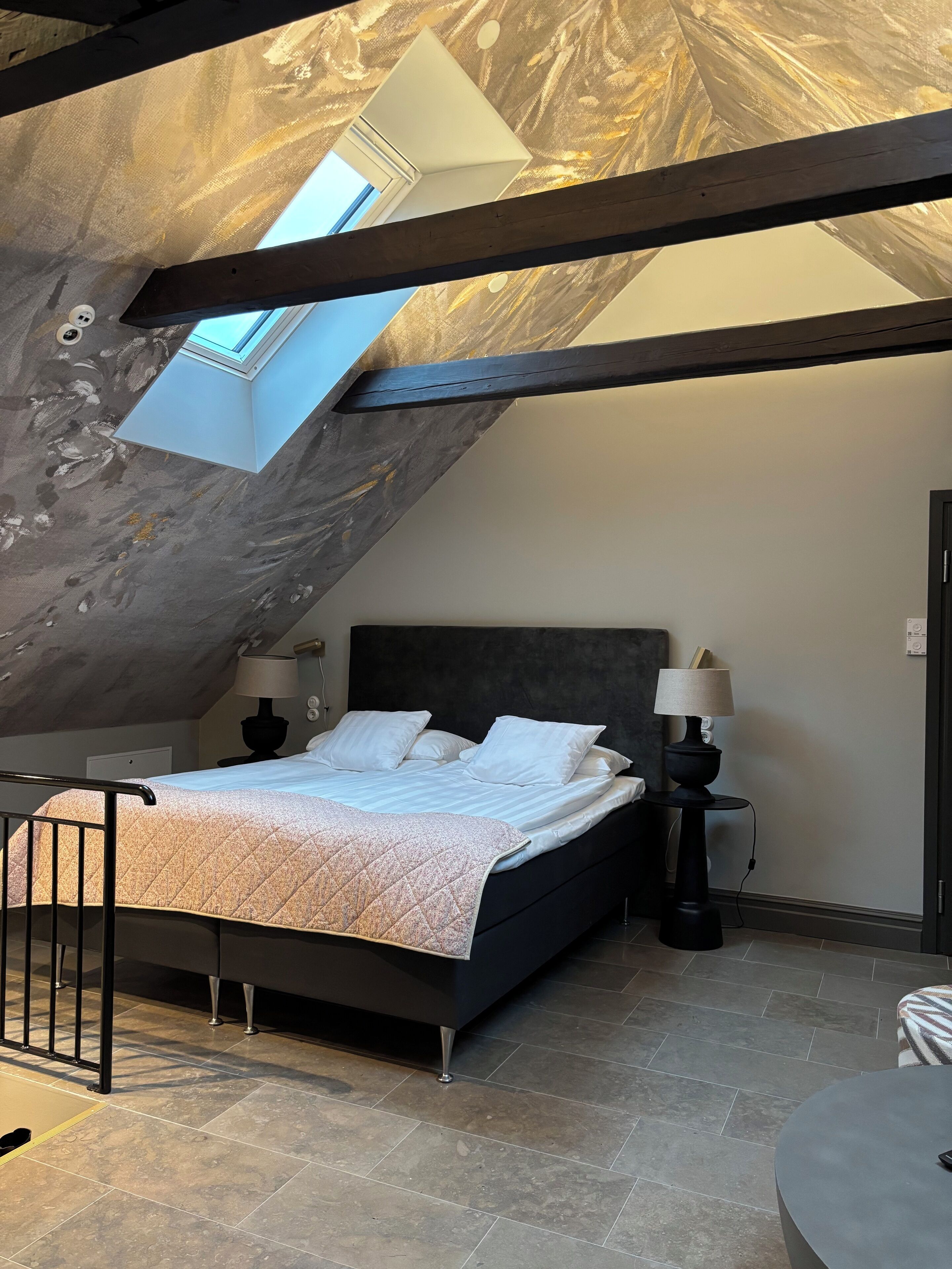 A bedroom with a sloped ceiling, a skylight, a bed with white bedding, two bedside lamps, and a small round table.