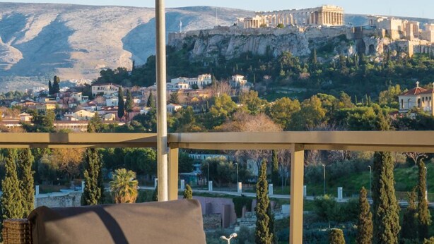 A wicker lounge chair with a cushion, overlooking a cityscape with ancient ruins on a hill. - Virgo Loft  Spectacular Acropolis View (Athens)