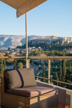 A wicker lounge chair with a cushion, overlooking a cityscape with ancient ruins on a hill. - Virgo Loft  Spectacular Acropolis View (Athens)