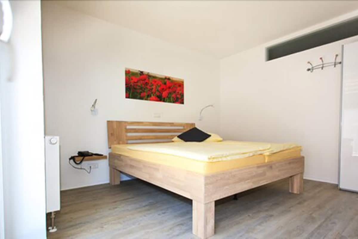 A bedroom with a wooden bed, a small bedside table, a wall-mounted light, and a door with a handle.