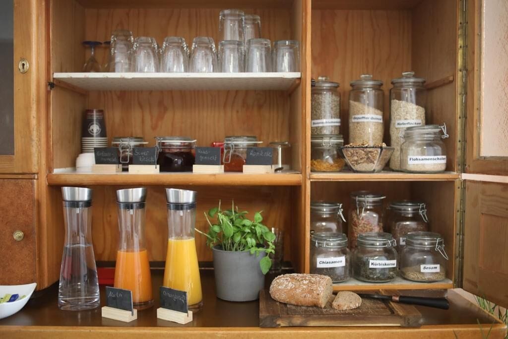 A kitchen area with shelves containing glass jars of various ingredients, a cutting board with a knife, and a potted plant.
