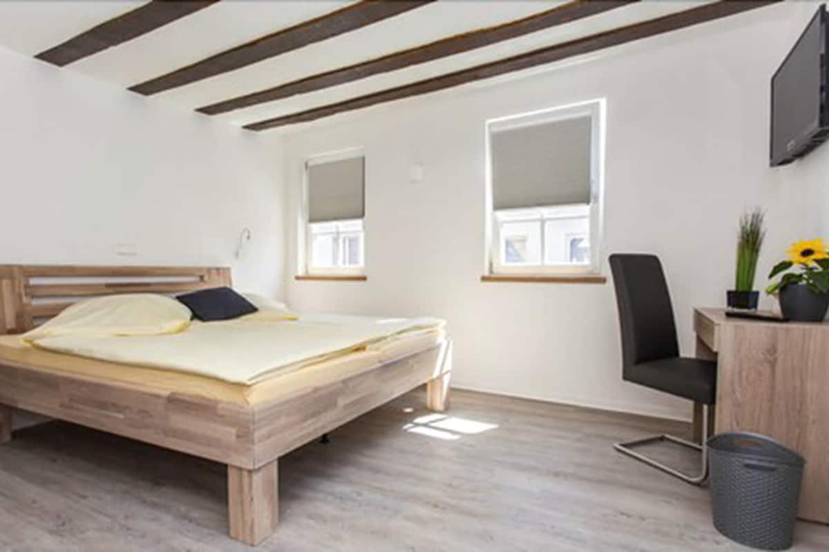 A bedroom with a wooden bed, a desk with a chair, a TV, and a window with a plant.