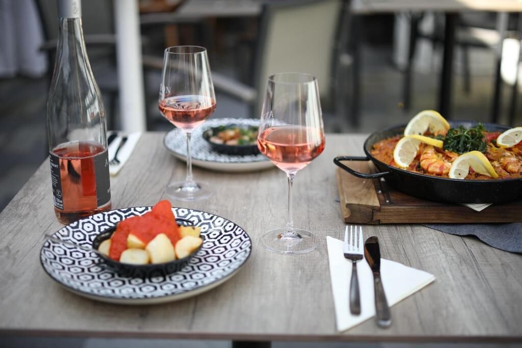 Una mesa de comedor con una botella de vino tinto, dos copas de vino, un plato con comida y una sartén con un guiso decorado con rodajas de limón.