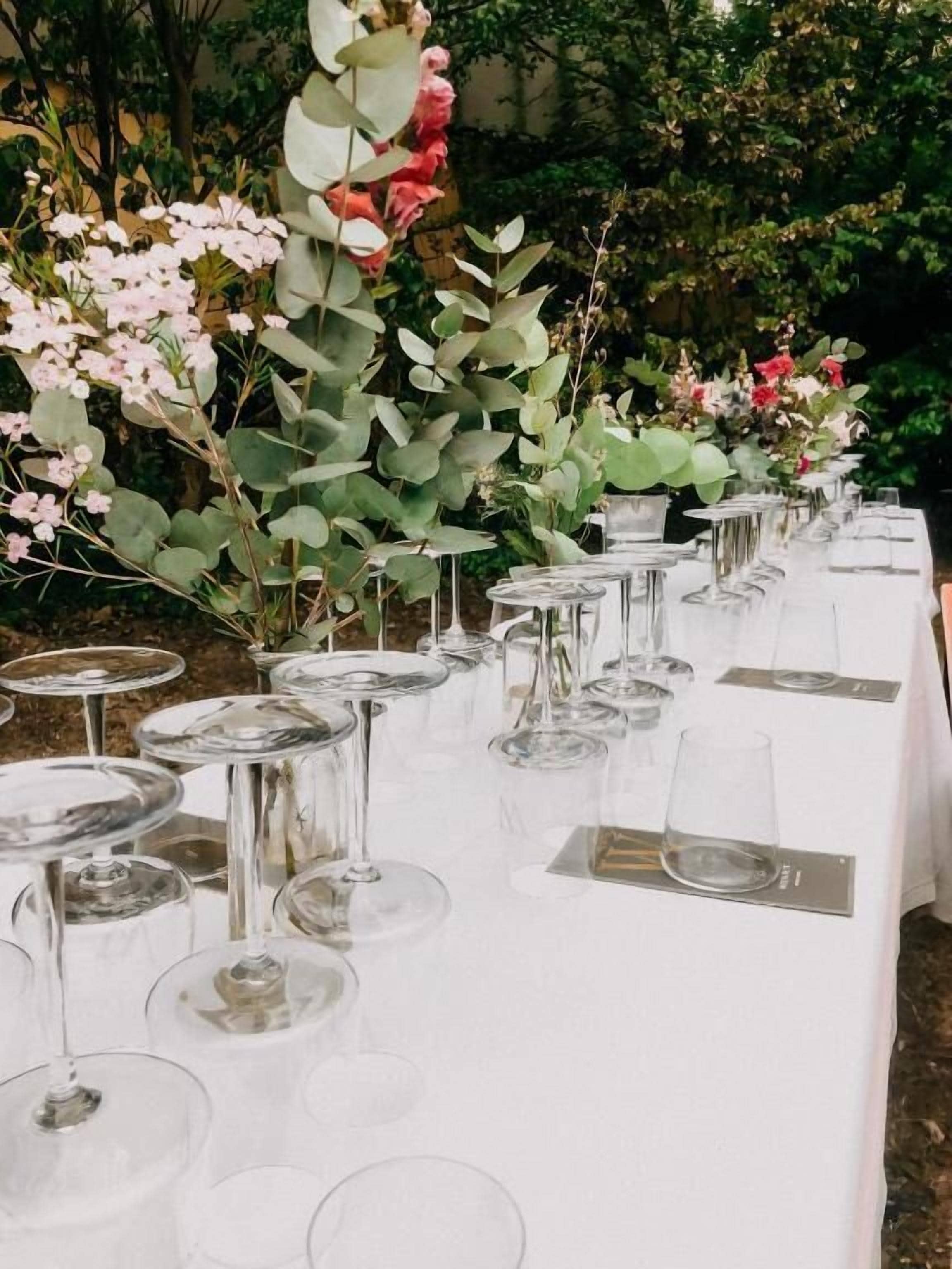 Una larga mesa dispuesta con copas de vino y platos, rodeada de follaje y flores.