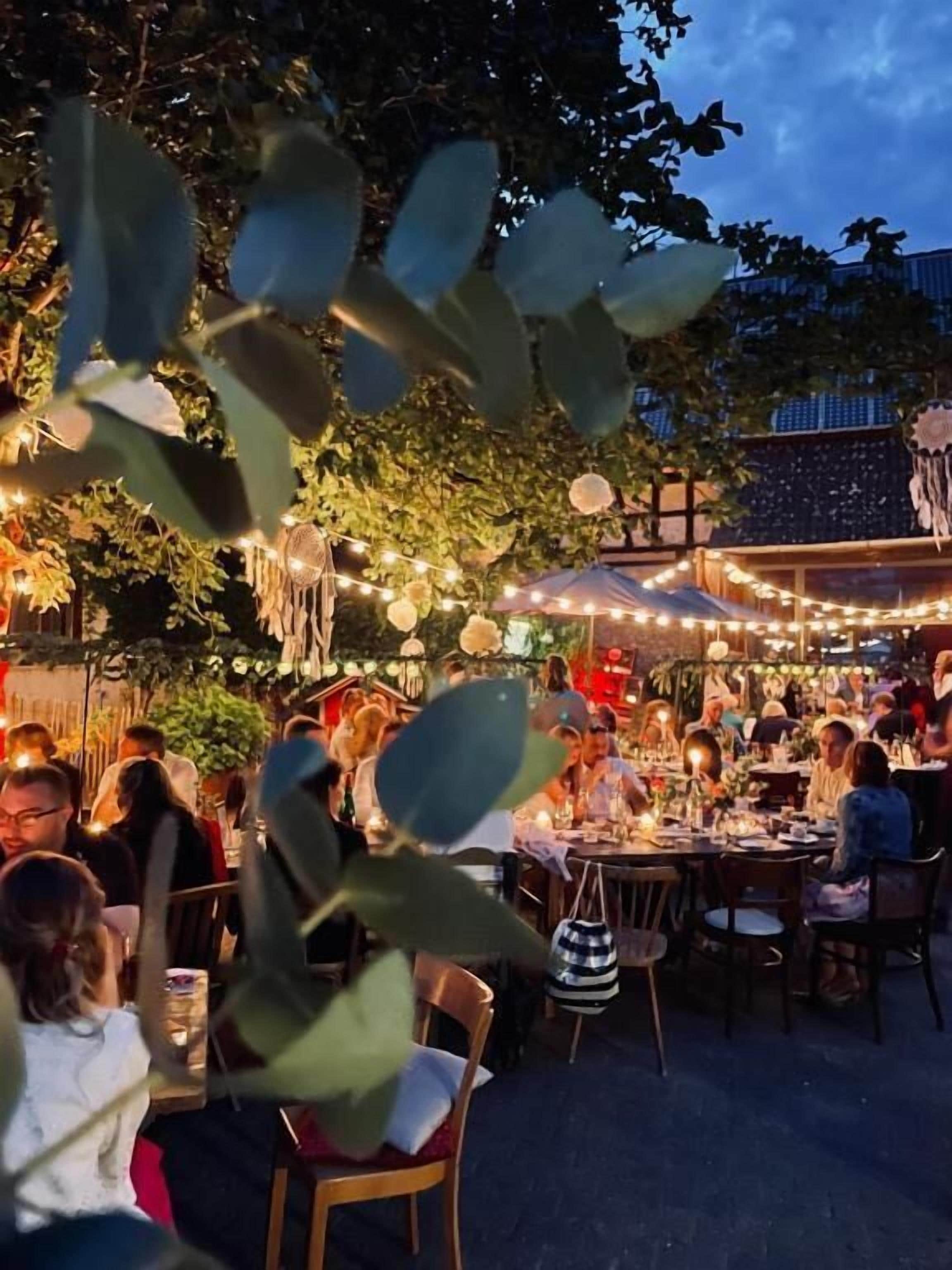 An outdoor dining area with tables and chairs, surrounded by foliage and string lights.