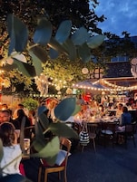 An outdoor dining area with tables and chairs, surrounded by foliage and string lights.
