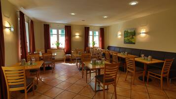 A restaurant with multiple wooden tables and chairs, a large window with red curtains, and wall-mounted lighting.