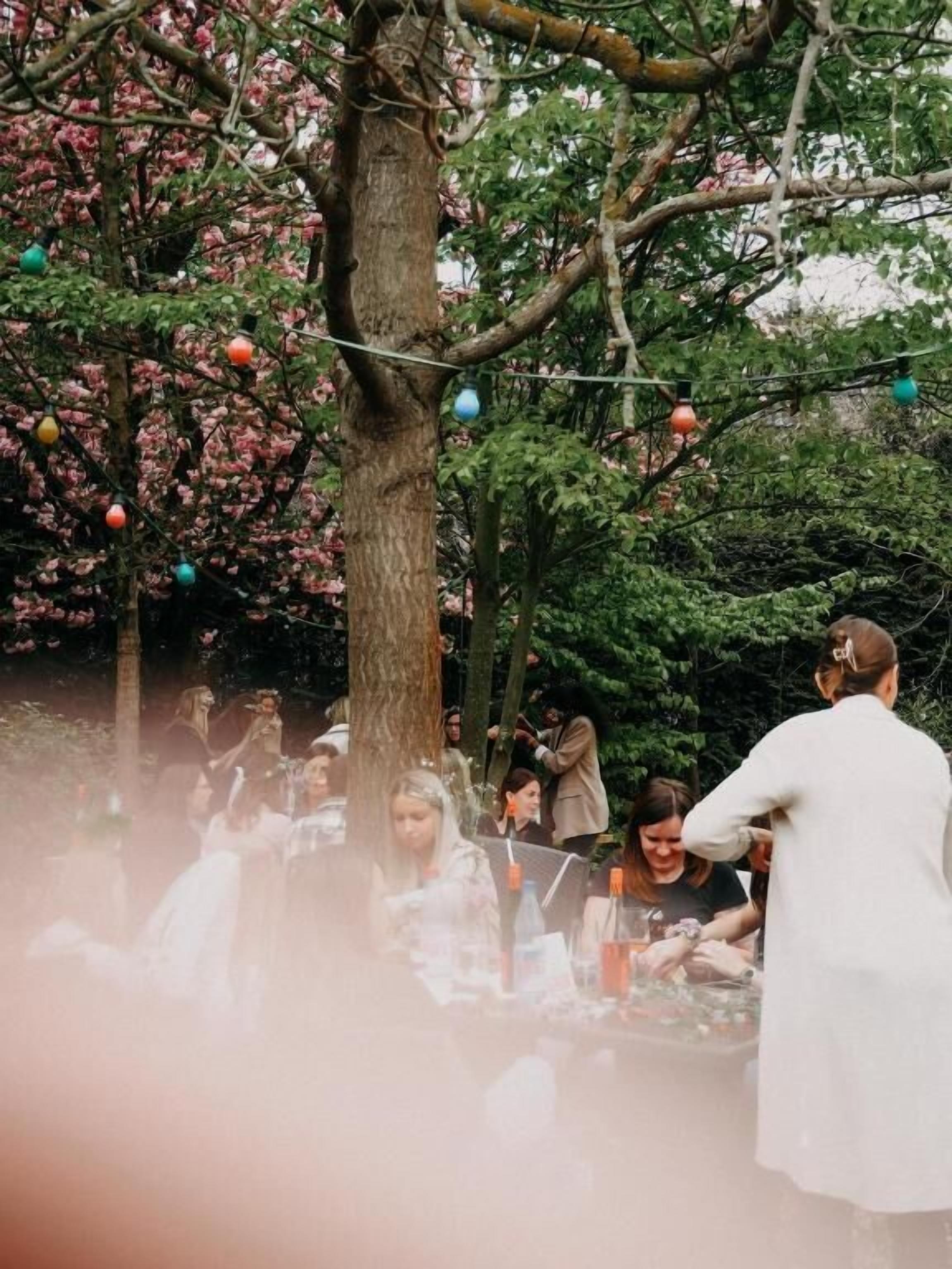 Eine Gruppe von Personen sitzt an einem Tisch im Freien, im Hintergrund ist ein Baum zu sehen.