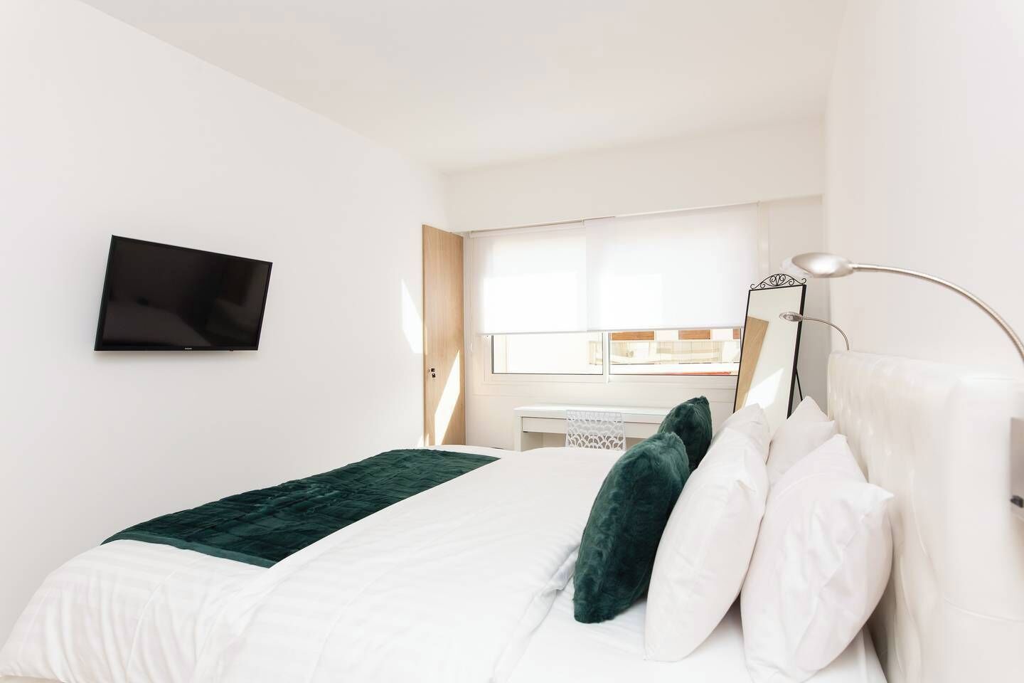 A bedroom with a bed, a TV mounted on the wall, a desk with a chair, and a window with blinds.