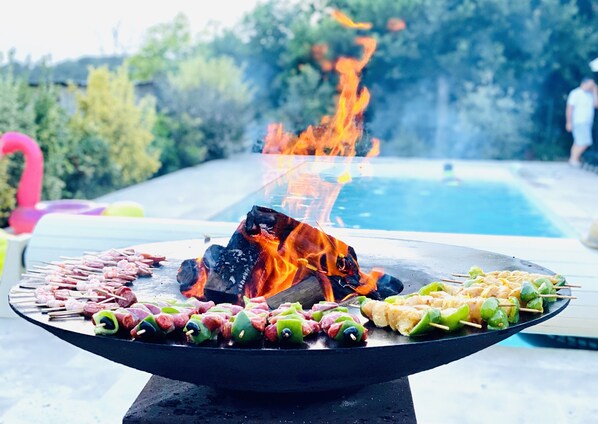 A barbecue grill with skewers of meat and vegetables, a pool, and a flaming fire in the background.